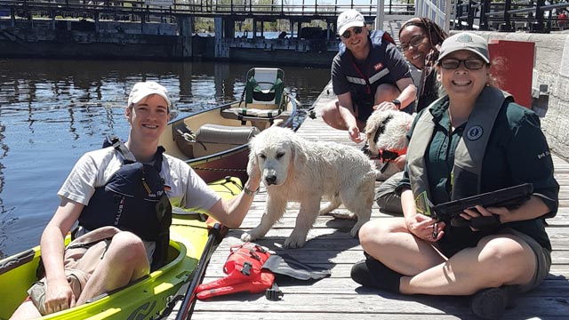 On a dock along the Lachine Canal, three people wearing life jackets sit near a kayaker, accompanied by dogs.