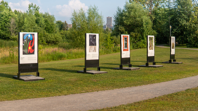 A view of five panels displaying the works of visual artists as part of an exhibition at the Cartier-Brébeuf national historic site.