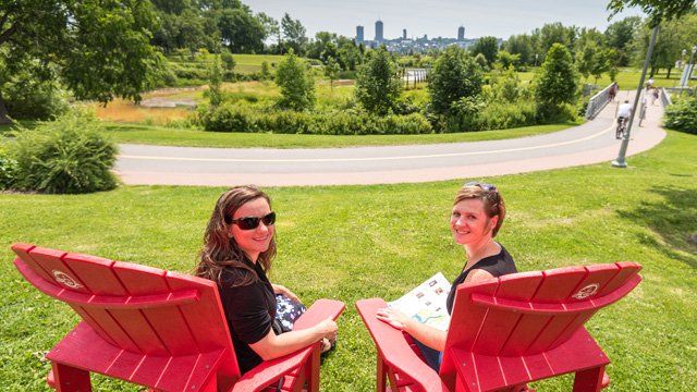 Vue de la piste cyclable à Cartier-Brébeuf et de deux femmes assissent sur de chaises rouges qui regardent vers la caméra.