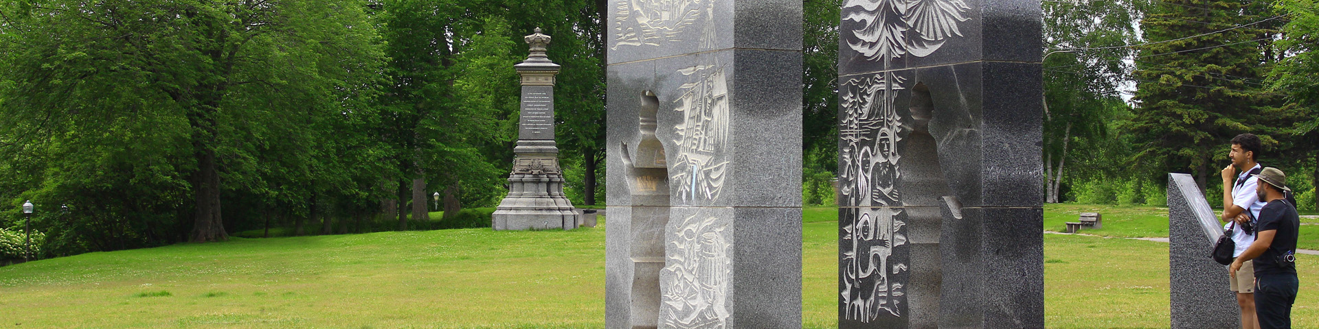 Visiteurs regardant un panneau en pierre et des sculptures au lieu historique national Cartier-Brébeuf.