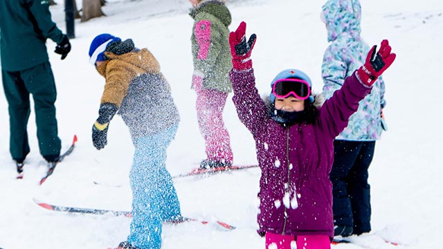 Children on skis playing in the snow; a young girl in the foreground raises her arms joyfully during an outdoor winter activity.
