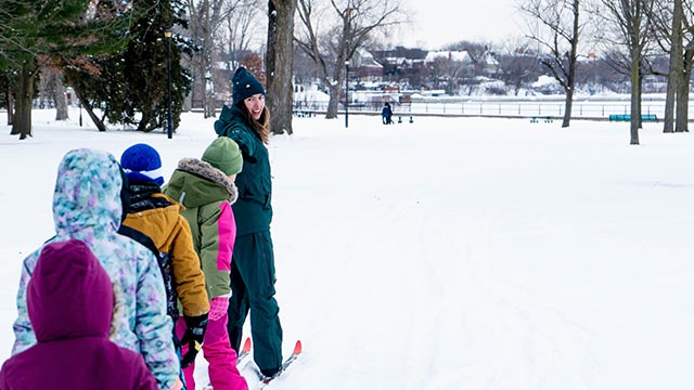 Group of children lined up on skis, wearing colorful winter clothing, following an instructor during a beginner ski activity in a snowy park.