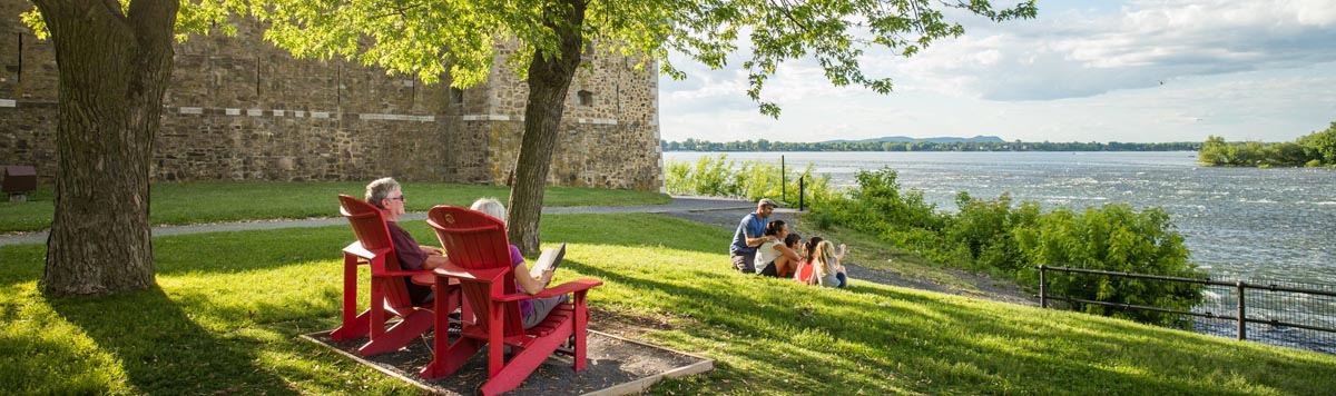 Aux abords du Fort, dans le parc du Fort Chambly, un couple lit sur des chaises rouges. Dans l’herbe, une famille observe la rivière Richelieu. Lieu historique national du Fort-Chambly. 