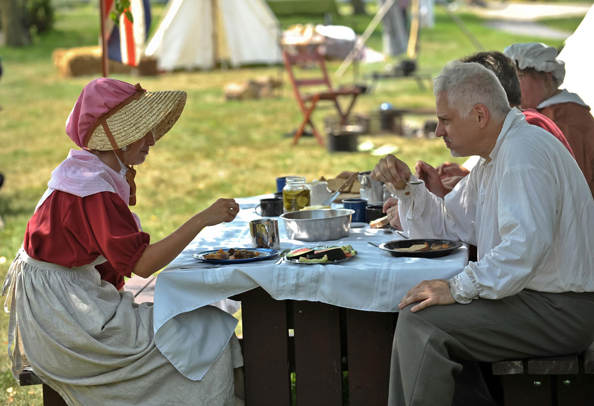 People in an era costume eating outside at a table.