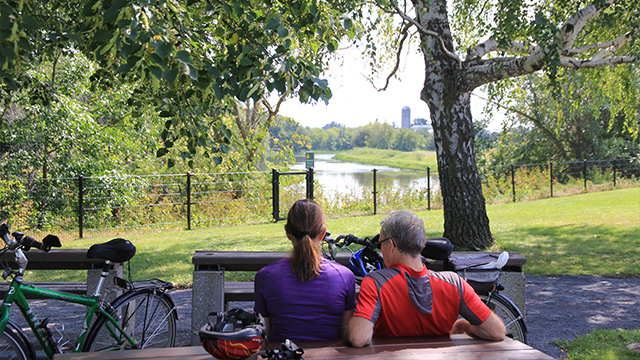 Deux cyclistes, un homme et une femme, font une pause sur un banc de parc au lieu historique national de la Bataille-de-la-Châteauguay et observent la rivière Châteauguay lors d’une journée d’été.