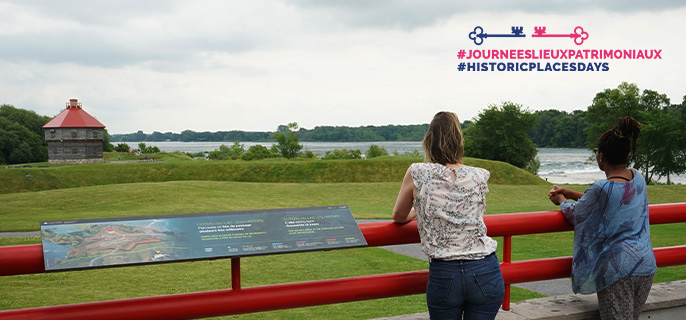 Deux visiteuses observent le blockhaus et la vue sur le fleuve Saint-Laurent lors d’une journée estivale au lieu historique national de Coteau-du-Lac.