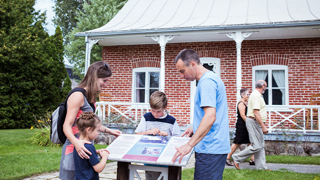 A father, a mother and their two children are looking at an interpretive panel in front of the house at Sir Wilfrid Laurier National Historic Site, while two elderly visitors walk nearby.