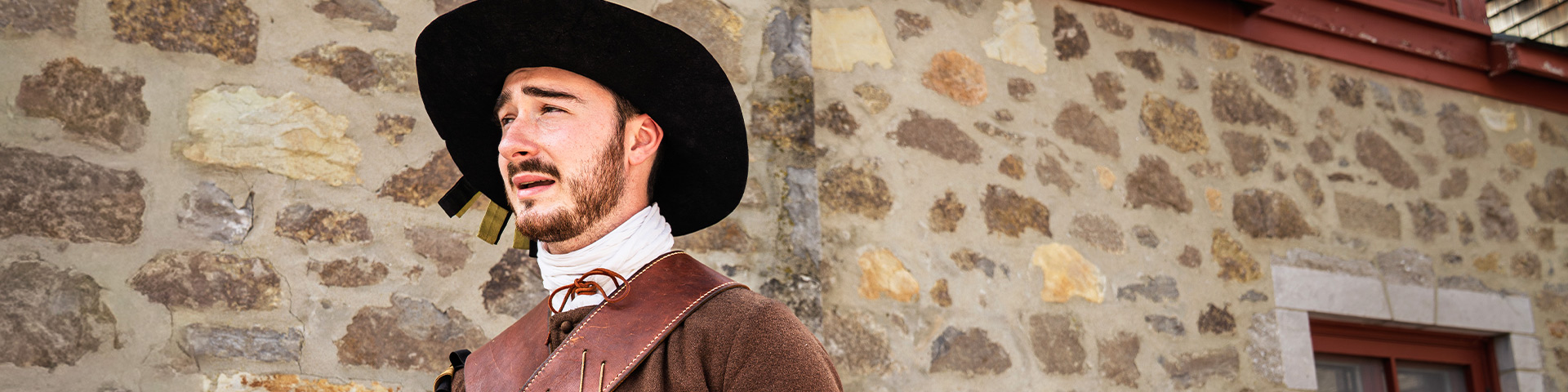 A Parks Canada guide, dressed as a soldier from the Carignan-Salières Regiment, stands in front of a stone wall in the open courtyard of Fort Chambly National Historic Site.