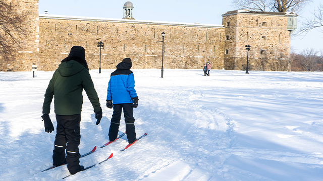 Two children learn cross-country skiing at Fort Chambly National Historic Site with Parks Canada guides, and the fort can be seen in the background against a winter landscape.