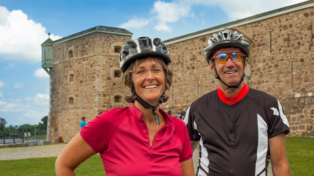 Two smiling cyclists, a man and a woman, pose in front of the Fort Chambly National Historic Site.