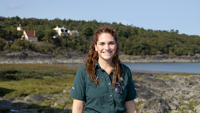A uniformed Parks Canada employee poses in front of the hospital bay with a view on the horizon of the Anglican chapel and a residence.