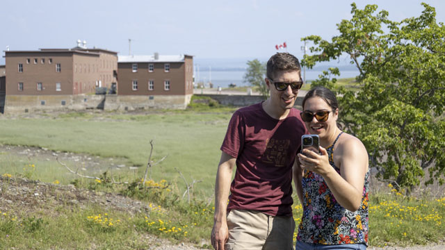 Deux visiteurs utilisent l'application de Parcs Canada via un téléphone portable dans le secteur ouest de Grosse-Île avec la baie de l'Hôpital et le bâtiment de désinfection en arrière-plan.