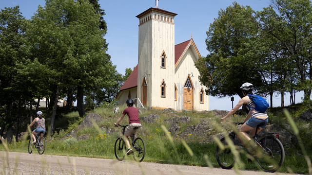 Three visitors ride bicycles in front of the Anglican chapel on the central path of Grosse Île.