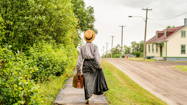  A young woman in a teacher's costume walks with her back to the camera on a concrete sidewalk towards a yellow building.