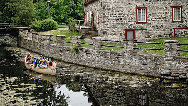 Deux guides de Parcs Canada animent une activité estivale de rabaska en groupe sur le canal de Lachine où on peut aussi apercevoir le lieu historique national du Commerce-de-la-Fourrure-à-Lachine.