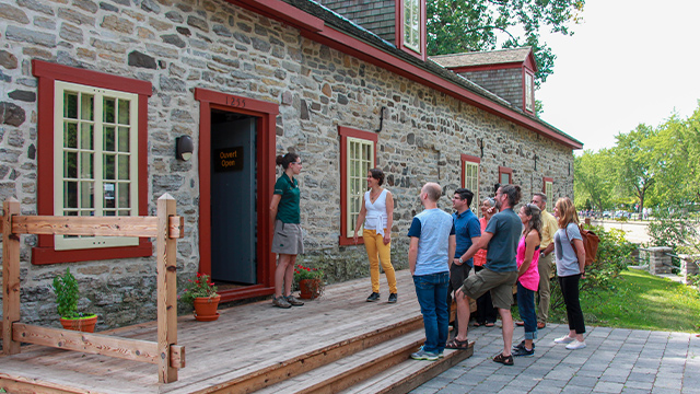Une guide de Parcs Canada, debout devant la porte de l’entrepôt de pierre, explique des faits historiques à un groupe de visiteurs adultes lors d’une visite au lieu historique national du Commerce-de-la-Fourrure-à-Lachine.