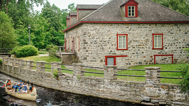 Un groupe de visiteurs sont dans un rabaska avec deux guides de Parcs Canada et ils naviguent sur le canal aux abords du lieu historique national du Commerce-de-la-Fourrure-à-Lachine.