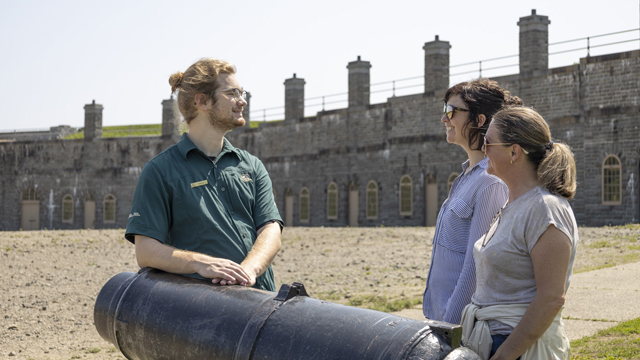  A Parks Canada guide leaning on a truck at the Lévis Forts National Historic Site gives explanations to two visitors with the casemates in the background.