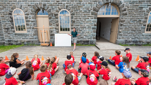 Une guide de Parcs Canada explique à un groupe d'enfant habillés en soldat britannique les consignes du jeu. 