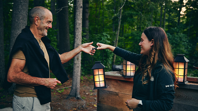 Une guide tend une lanterne à un couple de visiteurs âgés lors d'une visite guidée nocturne au lieu historique national du Manoir-Papineau.