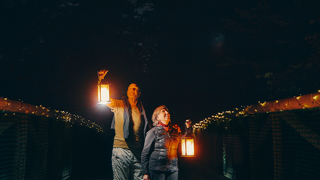 Two elderly people are visiting the Manoir Papineau National Historic Site at dusk, using lanterns for light.