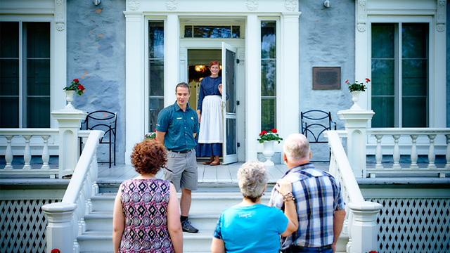 Three visitors, one man and two women, meet a character dressed as a gardener and a guide at the main entrance to the Manoir Papineau National Historic Site.