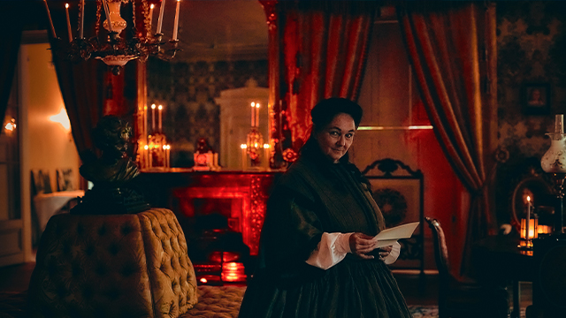 A female Parks Canada guide is dressed in a 19th-century bourgeois gown and is holding a book in the candlelit yellow lounge of the Papineau manor.