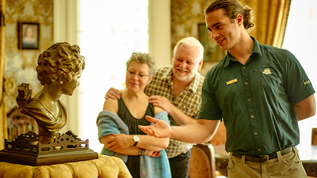 Un jeune guide de Parcs Canada explique à deux visiteurs âgés les particularités d’une statue exposée dans le somptueux salon jaune du lieu historique national du Manoir-Papineau.