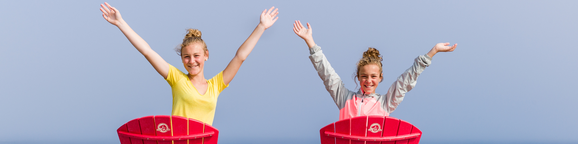 Deux fillettes assises sur des chaises rouges sourient à la caméra, les bras dans les airs.