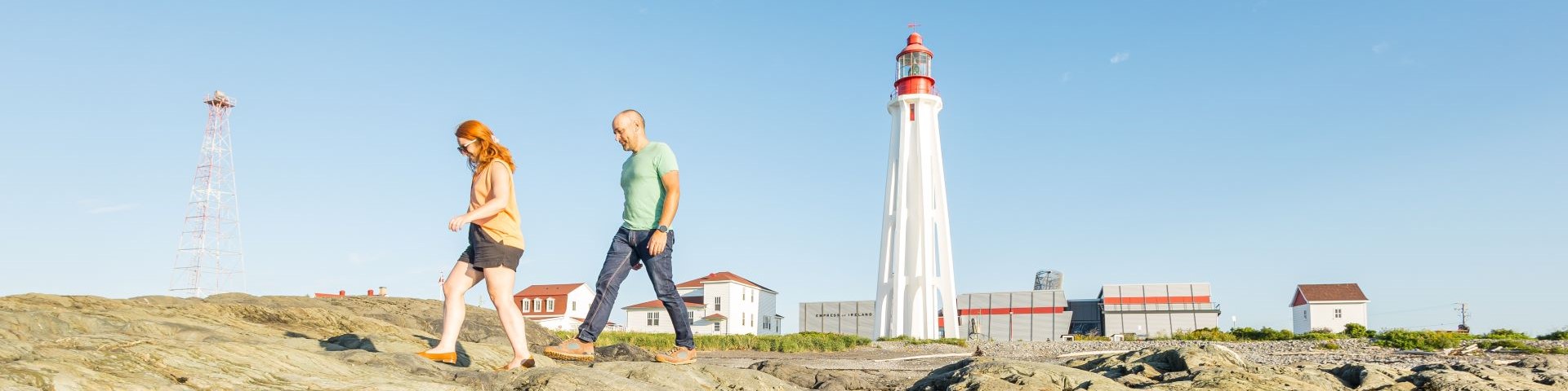 Two visitors are walking on a rocky shore in front a lighthouse and its dwellings. 
