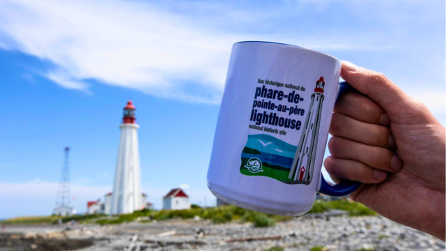 A mug of the Pointe-au-Père lighthouse held in front of the lighthouse station in background.