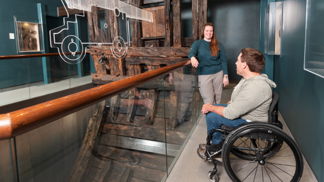 A visitor in a wheelchair chats with a Parks Canada employee in the exhibition room near a shipwreck. 