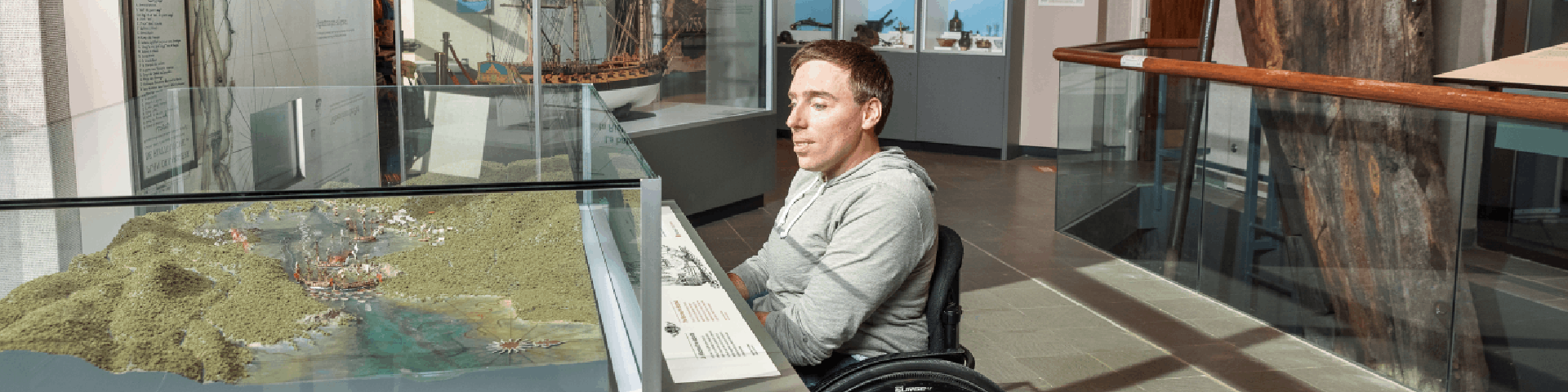 Un visiteur en fauteuil roulant regarde une scène de bataille navale miniaturisée dans un musée. 