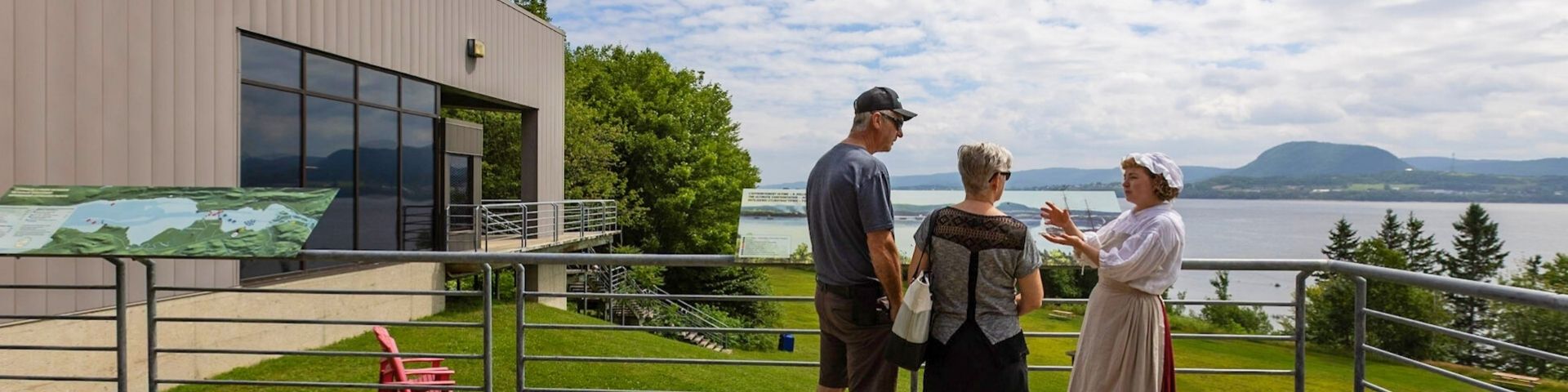 Une employée en costume d'époque discute avec deux visiteurs sur une terrasse près d'un musée. 