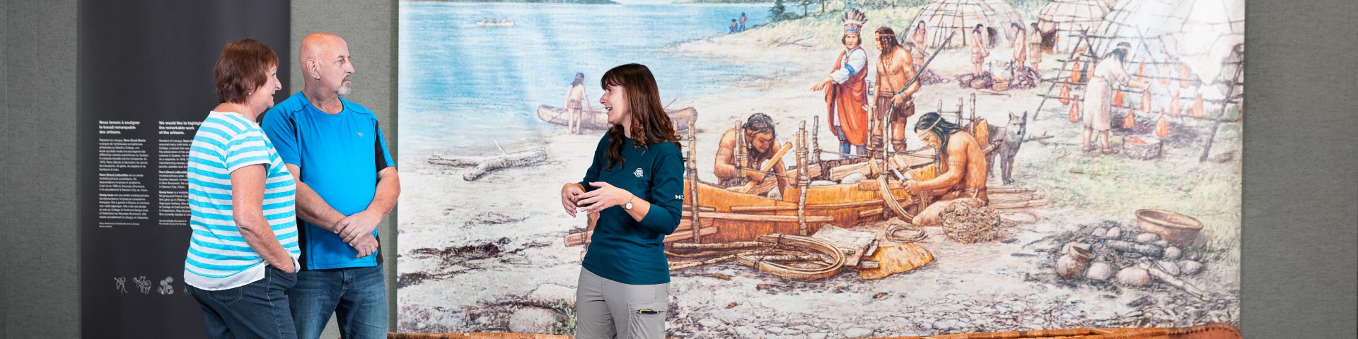 Deux visiteurs et une employée devant une fresque représentant une scène traditionnelle autochtone.