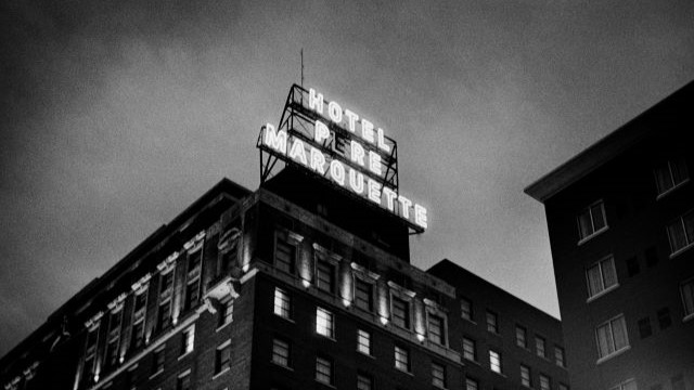 Photo en noir et blanc montrant la façade d'un immeuble avec un panneau lumineux sur le toit où on peut lire Motel Père Marquette. 