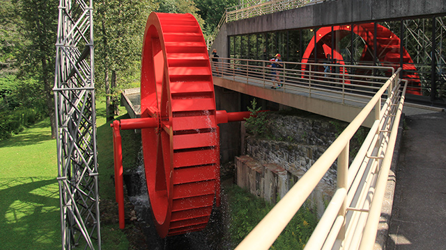 Des enfants accoudés sur la balustrade du haut fourneau, au lieu historique national des Forges-du-Saint-Maurice, regardent tourner la grande roue à eau de couleur rouge vif.