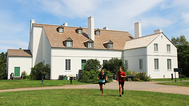 A boy and a girl are running and smiling on a beautiful summer day at Forges du Saint-Maurice National Historic Site, with the Ironmaster’s House in the background.