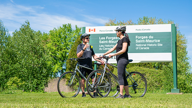 Un couple de cyclistes, un homme et une femme, se sont arrêtés à l’entrée du stationnement du lieu historique national des Forges-du-Saint-Maurice par une magnifique journée d’été; en arrière-plan, on aperçoit un grand panneau de signalisation de Parcs Canada affichant le nom du site, « Les Forges-du-Saint-Maurice ».