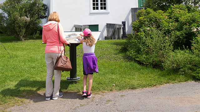 Photo of a mother and daughter visiting the Forges du Saint-Maurice National Historic Site, reading an interpretive panel outside the Grande Maison.