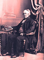 Un portrait de M. John McDougall assis dans un fauteuil près d'une table tenant un volume dans sa main droite. Il fut le propriétaire des Forges de 1863 à 1876.