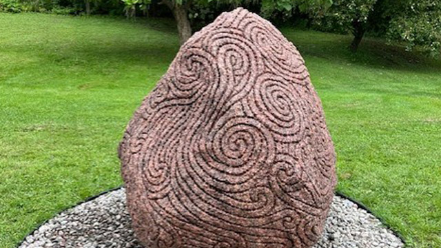 Une sculpture de pierre rougeâtre gravée de motifs en spirale et de lignes sinueuses est installée sur un lit de gravier circulaire, au milieu d’une pelouse verdoyante bordée d’arbres.