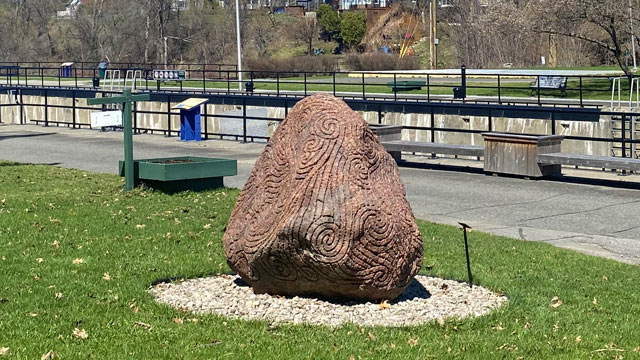 A reddish stone sculpture carved with spiral patterns and flowing, winding lines stands on a circular bed of gravel in the middle of a lush green lawn surrounded by trees.