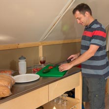 Famille dans une tente aménagée : un adulte prépare un repas pendant que des enfants jouent et se reposent sur des lits superposés, dans un espace chaleureux et fonctionnel.