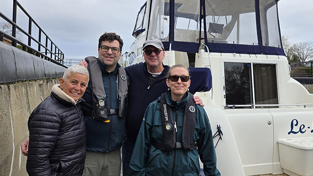 Quatre personnes se tiennent côte à côte devant un bateau blanc amarré, près d’un quai.