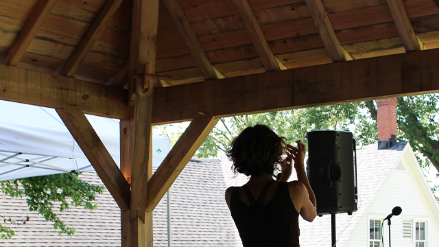 Une chanteuse est aperçue de dos et elle offre une prestation musicale sous le chapiteau en bois, appelé gloriette, au lieu historique national Louis-S.-St-Laurent; c’est l’été.