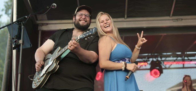 Le guitariste Marcus Quirion et la chanteuse Sandrine Hébert sont tous les deux très souriants sur scène lors d’un spectacle en plein air. La chanteuse, vêtue d’une robe bleue, fait un signe de paix avec les doigts, tandis que le guitariste joue avec enthousiasme.