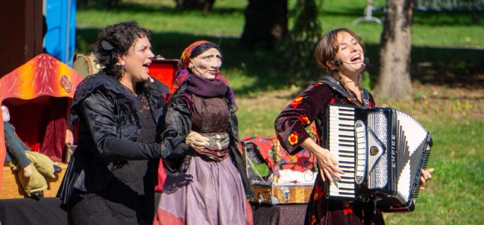 Un spectacle en plein air avec deux artistes sur scène. L'une des artistes joue de l'accordéon avec un grand sourire, tandis que l'autre manipule une marionnette, un personnage âgé vêtu d'un châle et d'un foulard. 