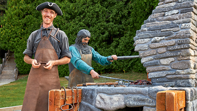 A smiling guide dressed as a blacksmith greets visitors near the outdoor carpentry and blacksmithing station at Obadjiwan–Fort Témiscamingue National Historic Site.