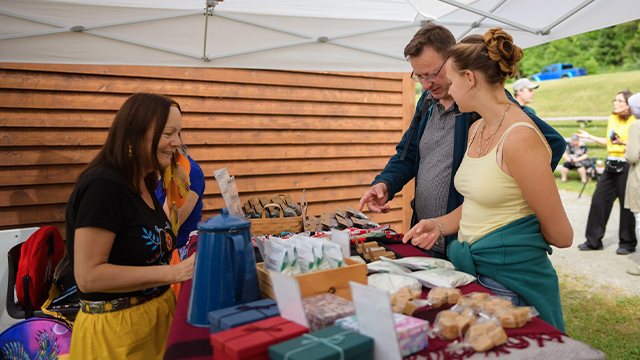 A man and a woman visiting Obadjiwan–Fort Témiscamingue National Historic Site stop by an outdoor Indigenous crafts kiosk to chat with an Algonquin woman who manages the sales.